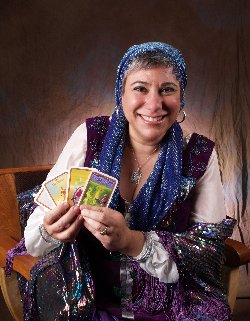 A smiling person wearing a blue headscarf and shiny scarf holds up four tarot cards while seated against a brown backdrop.